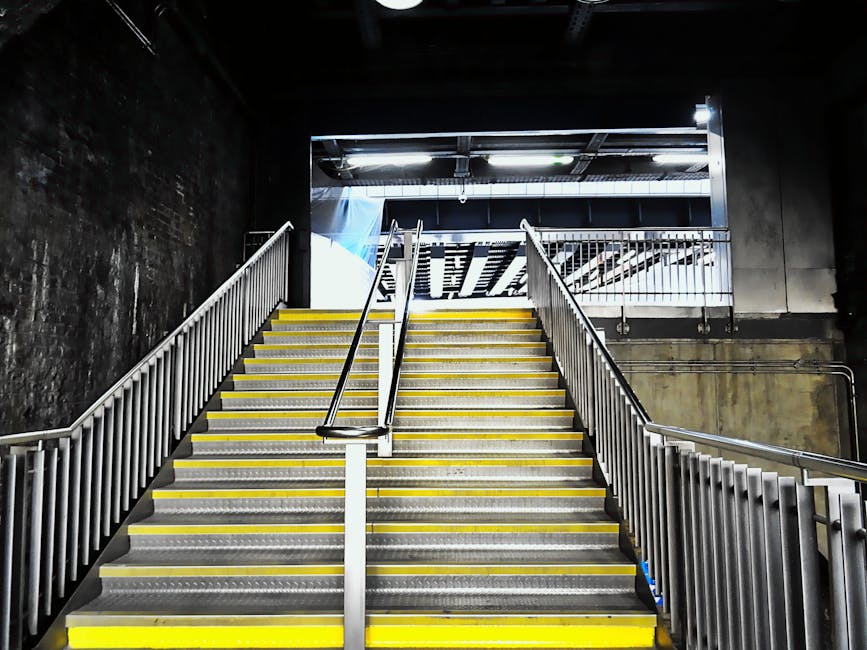 A wide shot of a metal staircase inside a building during a home relocation process, with safety yellow strips on the steps for improved visibility. The staircase features white metal railings on both sides, and the steps are made of metal with anti-slip surfaces. The surrounding environment includes dark, textured concrete walls and a ceiling with exposed piping, typical of an industrial or basement area. At the top of the stairs, an open doorway reveals a bright outdoor space or loading area, which is illuminated by natural light. The scene suggests a loading process where boxes, furniture, and packing materials could be carried up or down these stairs as part of a house removal service, with some packing materials, like plastic wrap and cardboard boxes, potentially visible nearby or nearby areas. Man and Van Southgate may be involved in navigating stair carries or access challenges during the move, using equipment such as trolleys or straps to facilitate safe transport of items through this stairway.