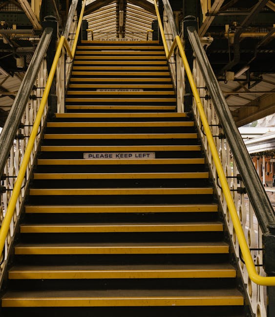 A wide set of industrial metal and wooden stairs with yellow handrails on both sides, leading upward within a warehouse or loading area. The stairs feature black strips on each step for traction, with a white sign in the middle reading 'PLEASE KEEP LEFT.' The surrounding environment includes structural metal beams and railing, indicating an indoor storage or logistics facility. Natural lighting illuminates the scene, highlighting the sturdy construction designed for moving or transporting furniture and boxes during a house removal or relocation process, as managed by Man and Van Southgate. The image emphasizes the practical aspects of navigating stairs in a commercial or residential move, reflecting careful planning in loading or unloading furniture and boxes, and ensuring safety during the home relocation activities.