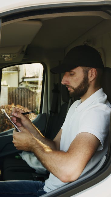 A man with a dark beard, wearing a black cap and a white polo shirt, sitting inside a delivery van and using a smartphone. The interior of the van has a light-colored ceiling and side panel, with a wi