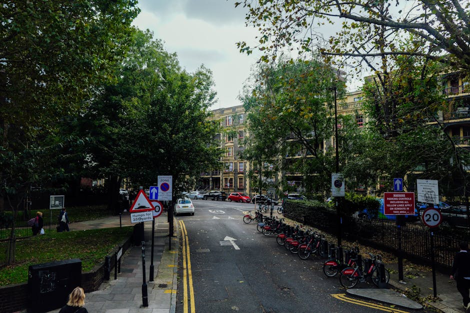 A street view in Southgate showing a tree-lined residential area with multi-storey apartment buildings in the background. In the foreground, there are several traffic and parking signs, including a triangular warning sign, a blue one-way sign, and a speed limit of 20 mph. To the right, a row of red and black rental bikes is parked along the pavement, near a fence. The street surface is dark asphalt with clearly marked yellow and white lines, and a white car is parked partially on the curb. On the left side, there is a sidewalk with pedestrians walking past, and some greenery and trees with dense foliage are visible along the edges of the street. The scene is overcast with diffused natural light, illustrating an urban environment suitable for house removals or furniture transport activities, supported subtly by signage and the street's layout.