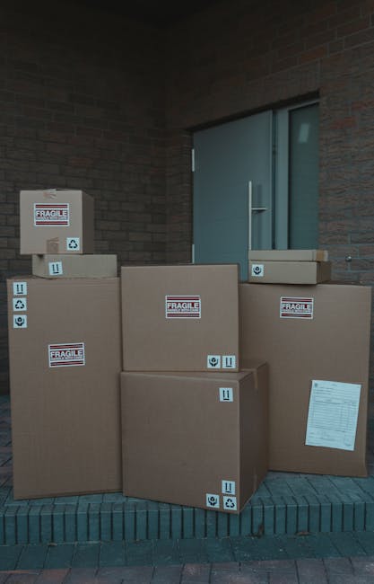 Several large cardboard boxes are stacked outside a building entrance, positioned on a paved area against a brick wall and a grey door. The boxes vary in size, with some placed directly on the ground and others leaned against the wall. Each box is labeled with a 'Fragile' sticker and standard handling symbols, indicating the contents require careful handling during a home relocation. A printed packing list is attached to one of the boxes. The boxes are secured with plastic straps, and the lighting suggests daytime with a neutral environment. This scene depicts the packing and loading process associated with house removals provided by Man and Van Southgate, highlighting efficient furniture transport and professional packing support during a move or relocation.