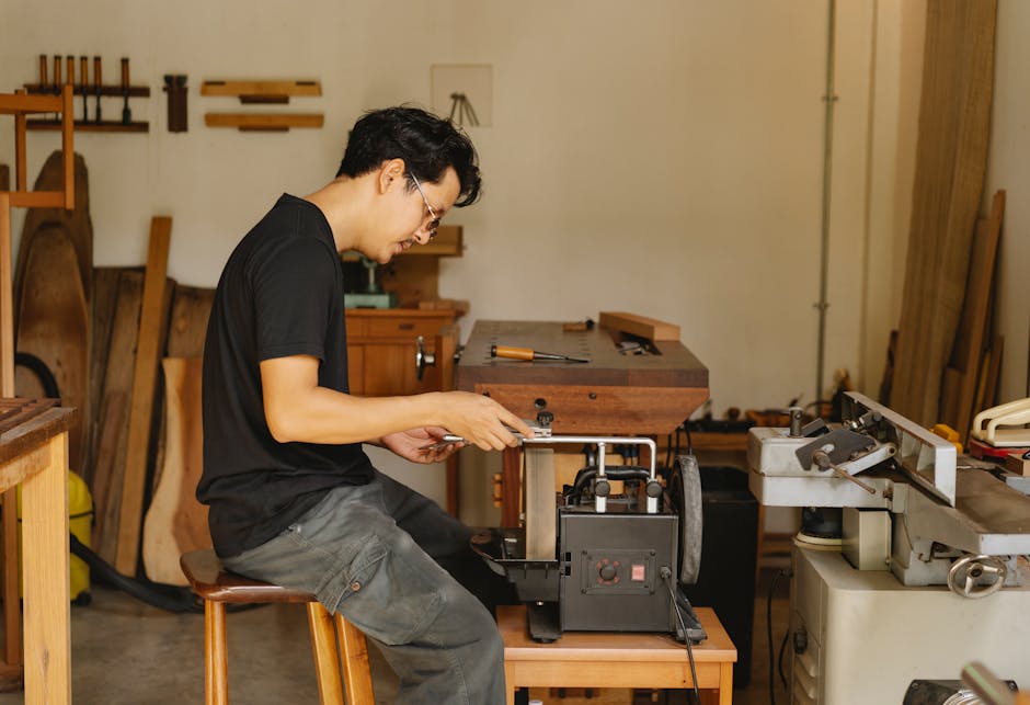 A man with dark hair, wearing glasses, a black t-shirt, and grey pants, is seated on a wooden stool inside a woodworking workshop. He is focused on operating a small piece of machinery, likely for furniture construction or repair. The workspace features various wooden objects, including a small wooden table or workbench, and wall-mounted tools such as saws and clamps. In the background, there are wooden planks and panels leaning against the wall, along with shelving units holding additional tools and materials. The lighting appears natural or softly diffused, illuminating the workspace evenly. This setting suggests home furniture crafting or repair, related to packing and preparing furniture for transport as part of a house removals process, which aligns with the services provided by Man and Van Southgate, experts in house removals and furniture transport.