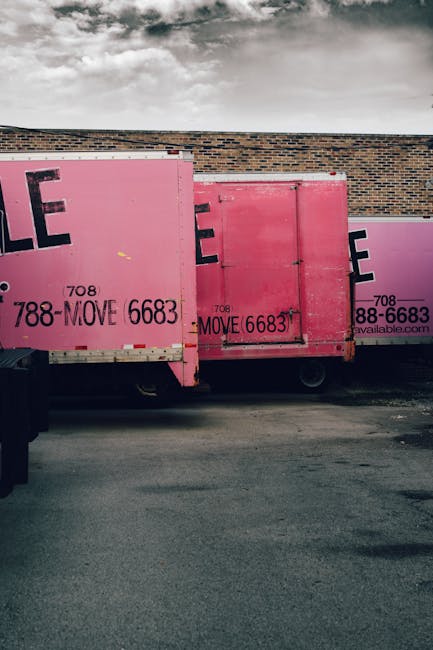 Several large cardboard boxes are stacked outside a building entrance, positioned on a paved area against a brick wall and a grey door. The boxes vary in size, with some placed directly on the ground and others leaned against the wall. Each box is labeled with a 'Fragile' sticker and standard handling symbols, indicating the contents require careful handling during a home relocation. A printed packing list is attached to one of the boxes. The boxes are secured with plastic straps, and the lighting suggests daytime with a neutral environment. This scene depicts the packing and loading process associated with house removals provided by Man and Van Southgate, highlighting efficient furniture transport and professional packing support during a move or relocation.