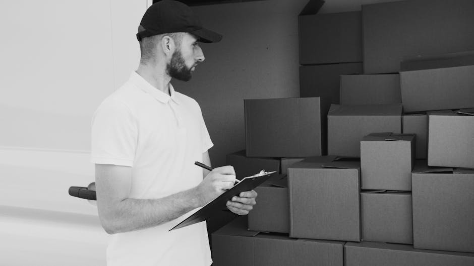 A man with a beard, wearing a white polo shirt and a black cap, is standing indoors beside a stack of uniform cardboard boxes arranged on the floor against a plain wall. He is holding a clipboard and pen, appearing to take inventory or check off items as part of the packing process during a home relocation. The boxes vary in size and are packed with packing materials such as plastic wrap and paper, indicating careful preparation for a furniture transport or moving service. Behind him, a doorway leads into another room, and natural light illuminates the scene, highlighting the organized packing area. This setting demonstrates the loading and packing phases typical of professional removals, with a focus on accurate inventory management, which is essential to avoid common booking mistakes with Southgate man and van services, such as missing items or incorrect packing. Man and Van Southgate supports efficient furniture transport and moving logistics, ensuring a smooth home relocation process.