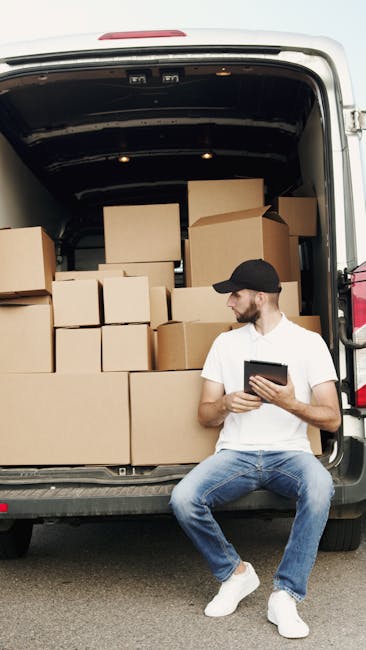 A man with a beard, wearing a white polo shirt and a black cap, is standing indoors beside a stack of uniform cardboard boxes arranged on the floor against a plain wall. He is holding a clipboard and pen, appearing to take inventory or check off items as part of the packing process during a home relocation. The boxes vary in size and are packed with packing materials such as plastic wrap and paper, indicating careful preparation for a furniture transport or moving service. Behind him, a doorway leads into another room, and natural light illuminates the scene, highlighting the organized packing area. This setting demonstrates the loading and packing phases typical of professional removals, with a focus on accurate inventory management, which is essential to avoid common booking mistakes with Southgate man and van services, such as missing items or incorrect packing. Man and Van Southgate supports efficient furniture transport and moving logistics, ensuring a smooth home relocation process.