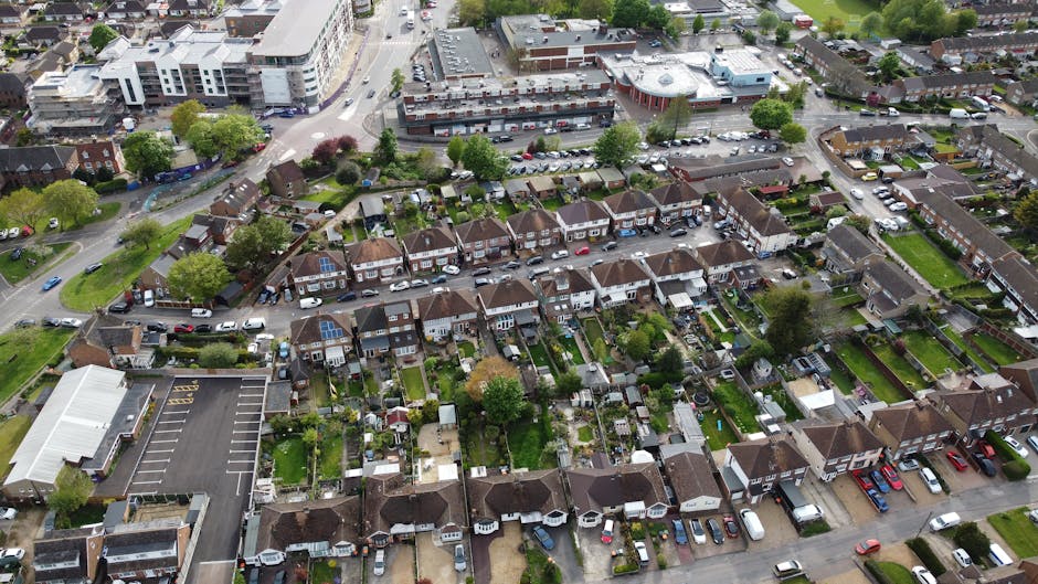 An aerial view of a residential neighbourhood showing rows of terraced houses with small backyards and gardens, lined with parked cars along the roads and driveways. Some houses have solar panels installed on their roofs, and a few garden features like sheds and patios are visible. The street is lined with trees and green spaces, with a mix of commercial and residential buildings in the background. The scene captures a typical suburban area suitable for home relocation and furniture transport, with residential property access points close to the pavement and parking areas nearby, reflecting the logistics involved in house moves managed by Man and Van Southgate.