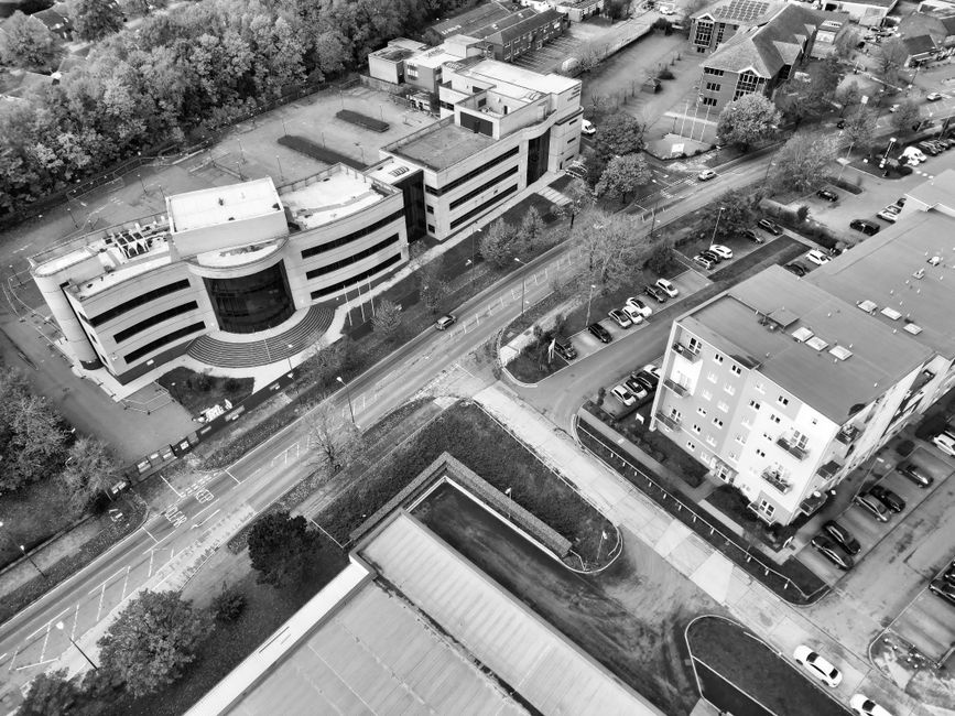 A black-and-white aerial photograph showing a multi-storey commercial building with large curved windows and a circular entrance, surrounded by trees and open green spaces. Adjacent to the building is a parking lot with multiple parked cars, and a street with moving vehicles. In the foreground, there is a rectangular covered structure, possibly a bus shelter or loading bay, alongside a pathway leading to the building. Nearby, a residential block with multiple floors and balconies is visible, with cars parked along the adjacent street and in designated parking areas. The scene captures the environment during daylight, with shadows indicating the light source. Man and Van Southgate uses this type of imagery in their service pages related to house removals, logistics, and home relocation to visually depict moving environments and urban settings for furniture transport and packing and moving processes.