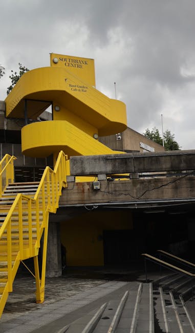 The image depicts a multi-level parking structure with a notable bright yellow, spiral exterior staircase leading to an upper floor. The building is part of the Southbank Centre, as indicated by a sign at the top of the stairwell. The concrete parking area below is visible, with metal ramps and a sloped walkway. Surrounding trees and a cloudy sky are seen in the background. This scene showcases the architectural design of a busy urban venue, with accessibility features such as the prominent staircase. The setting is outdoors, and the lighting appears natural under an overcast sky. In the context of house removals or furniture transport, this image may relate to loading or unloading activities at a site with distinctive architectural features, illustrating the logistical considerations of moving objects in urban environments. Man and Van Southgate's services could include handling such relocation projects involving complex access points or multi-storey structures.