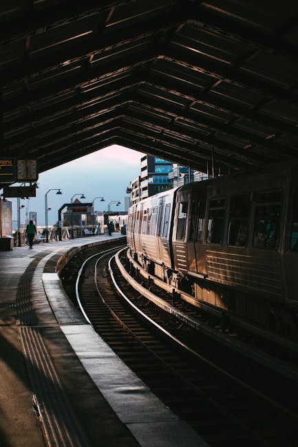 The image shows a train station platform viewed from inside a sheltered area with a high, triangular metal roof structure. On the platform, there are several cardboard boxes and plastic-wrapped furniture items arranged for transport, some of which are being carried or prepared for loading. A worker, dressed in casual clothing, is seen handling the packing materials, possibly during a home relocation or moving process. To the right, a modern metal train is stationary on the tracks, with windows reflecting ambient light. In the background, a few passengers are walking along the platform, and the station's entrance and exterior can be seen through the open edge of the shelter, with some streetlights and buildings further beyond. This scene exemplifies the logistics involved in furniture transport and packing for moving services such as those offered by Man and Van Southgate, particularly during the loading phase of a house removal near Southgate Station.