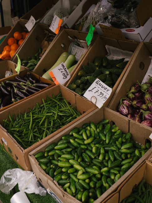 A display of fresh vegetables at a market, with multiple open cardboard boxes arranged on a table or stall. The boxes contain cucumbers, green beans, and purple-striped eggplants. Some of the vegetabl