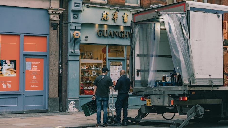 Two men, one dressed in a black t-shirt and jeans and the other in a dark jacket and trousers, are preparing to load a delivery truck parked outside a building with a grey facade and an Asian-themed storefront. The man in the black t-shirt is holding a shopping basket, while the other man appears to be guiding or communicating. The truck's back door is open, revealing a plastic curtain and some packaging materials inside. The scene takes place on a pavement in front of the shop, which features a large display window with items and notices, and gold Chinese characters above the entrance. The backdrop includes a blue door to the left and a partial view of adjacent buildings. The activity suggests a home or business relocation, involving furniture and boxed items being prepared for transportation, typical of a professional removals process handled by [COMPANY_NAME], fitting the context of same-day removals delays and moving logistics in Southgate.