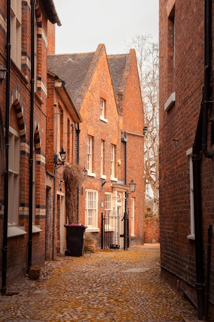 A narrow cobblestone alleyway situated between red brick residential buildings, with a black wrought iron gate at the end and a black wheelie bin positioned near the left side. The buildings feature traditional sash windows with white frames, some with small light fixtures mounted on the walls. Overhanging ivy or climbing plants are visible on one of the buildings, and leafless trees cast shadows on the scene, indicating an autumn or winter setting. The lighting is natural and slightly diffused, typical of an overcast day. This scene exemplifies the tight urban environment often encountered during house removals or furniture transport in historic housing areas, where careful navigation and logistical planning are required for successful move coordination, relevant to the Southgate N14 removals guide for narrow streets. Man and Van Southgate, a specialist in removals, likely manages such residential moves involving external access and precise handling of items through confined spaces.