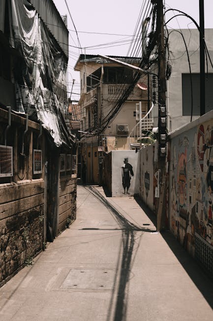 A narrow urban street with a concrete pavement lined by closely packed buildings on both sides. On the left, there are residential structures with worn brick walls, some covered with black plastic sheeting and makeshift renovations. To the right, a high wall is decorated with colorful graffiti art. Overhead, a tangle of exposed electrical wires and cables crisscross between utility poles, contributing to the cluttered cityscape. In the background, a person dressed in dark clothing is walking away down the street, partially silhouetted against the bright daylight. The street appears to be part of an older, densely populated neighbourhood, indicative of an area suitable for house removals and furniture transport services. The overall scene captures the typical environment where relocating items through narrow streets requires careful planning and logistical expertise, which [COMPANY_NAME] provides in house relocations and moving services, such as packing, loading, and transporting furniture and boxes in tight urban environments.