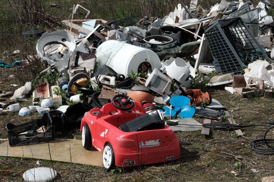 Outside a property, a large pile of discarded household appliances, furniture, and packaging materials is situated on the ground, indicating waste disposal during a home relocation or clearance process. The heap includes broken washing machines, televisions, plastic containers, cardboard boxes, and metal objects, with some items scattered across a patch of bare earth and greenery. A small red and black children's ride-on car is positioned in the foreground, showing signs of wear with scratches and chipped paint. Nearby, a black plastic crate and various tangled cables rest on the ground, while a wooden plank acts as a pathway leading toward the pile. The background features overgrown bushes, trees, and a partially visible pathway, suggesting the site is adjacent to a residential or semi-natural area. The scene is captured in daylight with natural lighting, reflecting a typical loading or clearing scene during household item disposal, guided by Man and Van Southgate's removals services focused on efficient waste removal and furniture transport.
