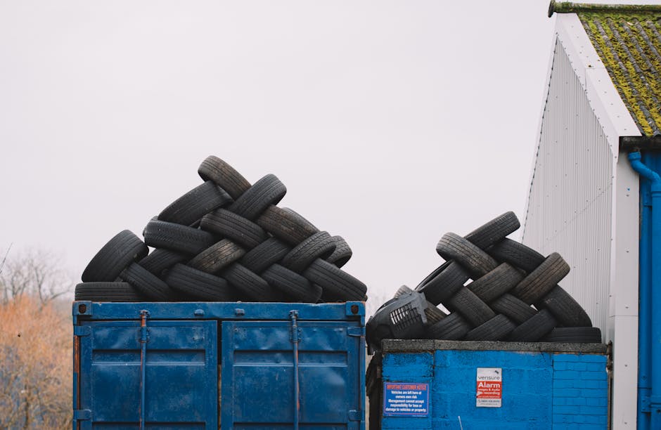 Two blue skip bins filled with numerous stacked black tyres in an outdoor area next to a white building with a blue pipe visible on the wall. One skip is positioned directly on the pavement, while the other is adjacent to the building, both containing tyres arranged in a haphazard manner. The tyres are of various sizes, with some appearing worn and others newer. The background shows a cloudy sky and some distant leafless trees, indicating a cold or overcast day. This image captures loading and disposal processes related to house removals or decluttering, which Man and Van Southgate may assist with during home relocation projects. The tyres symbolize waste or unwanted items that may need proper disposal or recycling during moving or clearance services. The environment is clean, with no other furniture or packaging materials visible, emphasizing the focus on tyre disposal within the context of property clearance and moving logistics.