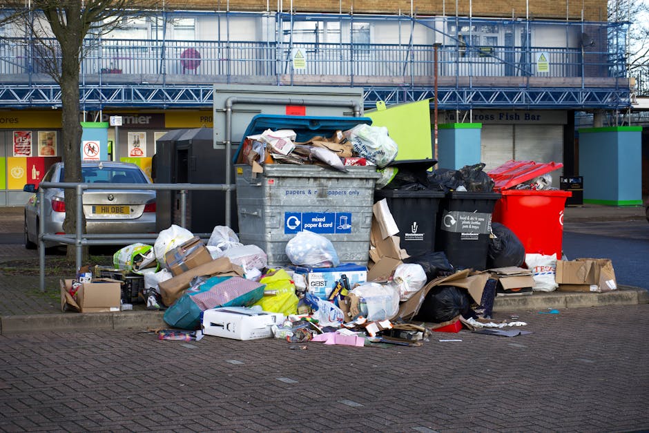 Outside a property, a large pile of discarded household appliances, furniture, and packaging materials is situated on the ground, indicating waste disposal during a home relocation or clearance process. The heap includes broken washing machines, televisions, plastic containers, cardboard boxes, and metal objects, with some items scattered across a patch of bare earth and greenery. A small red and black children's ride-on car is positioned in the foreground, showing signs of wear with scratches and chipped paint. Nearby, a black plastic crate and various tangled cables rest on the ground, while a wooden plank acts as a pathway leading toward the pile. The background features overgrown bushes, trees, and a partially visible pathway, suggesting the site is adjacent to a residential or semi-natural area. The scene is captured in daylight with natural lighting, reflecting a typical loading or clearing scene during household item disposal, guided by Man and Van Southgate's removals services focused on efficient waste removal and furniture transport.